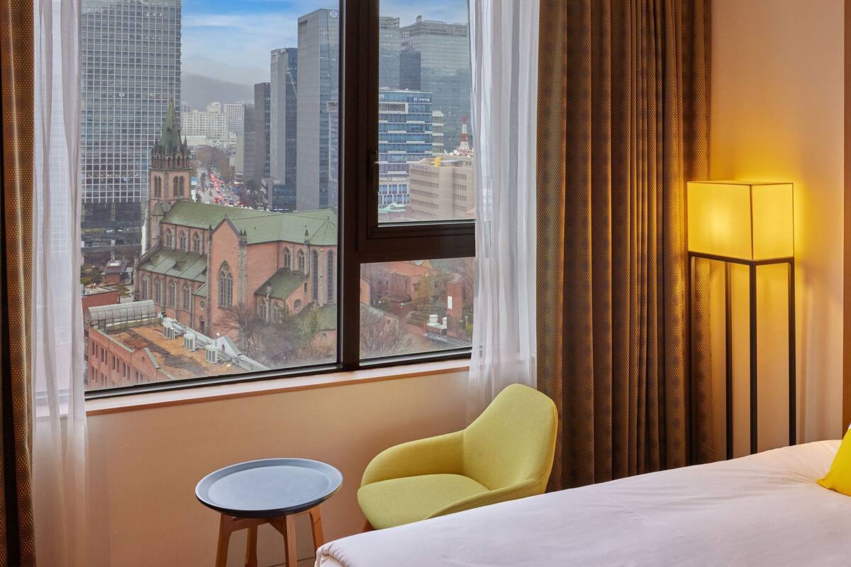 Hotelkamer met een bed, een stoel, een klein tafeltje, een lamp en uitzicht op een stadsbeeld door een groot raam.