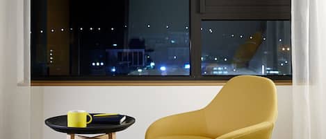 A hotel room with a bed, a yellow armchair, a small table with a mug, and a view of a cityscape with a tower at night.