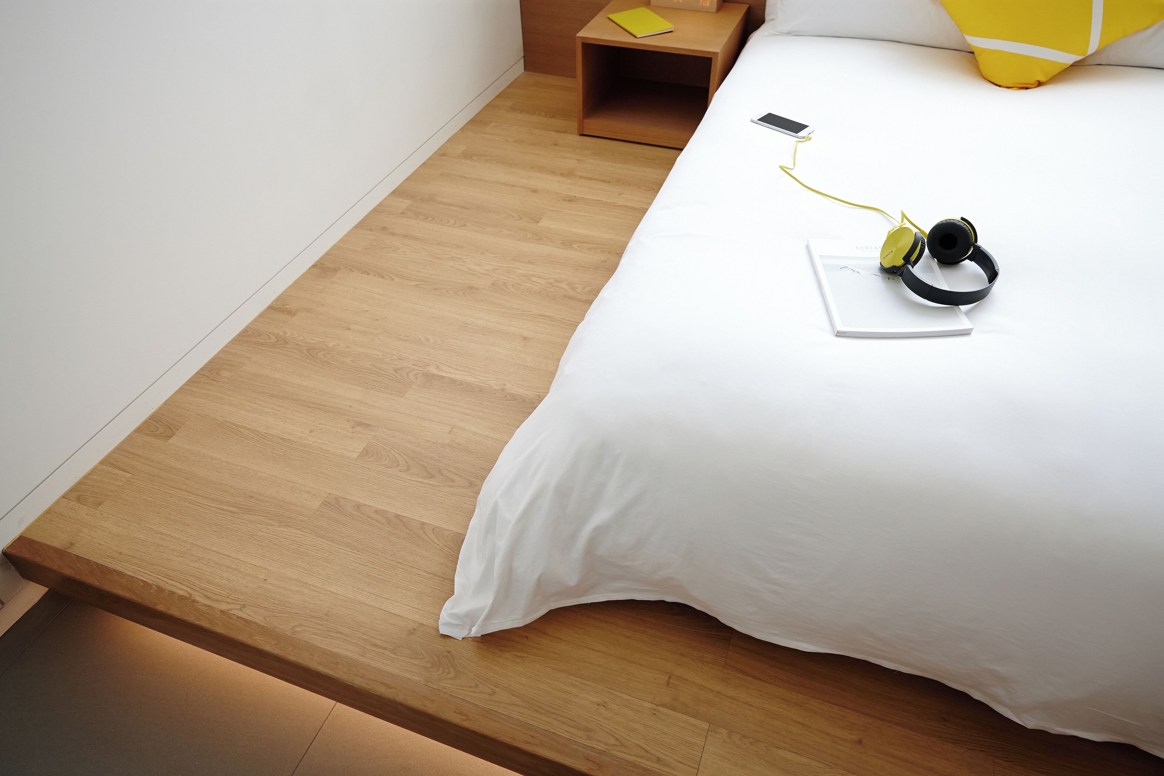 A neatly made bed with a white sheet, a wooden nightstand with a yellow book, and a pair of headphones on the bed.