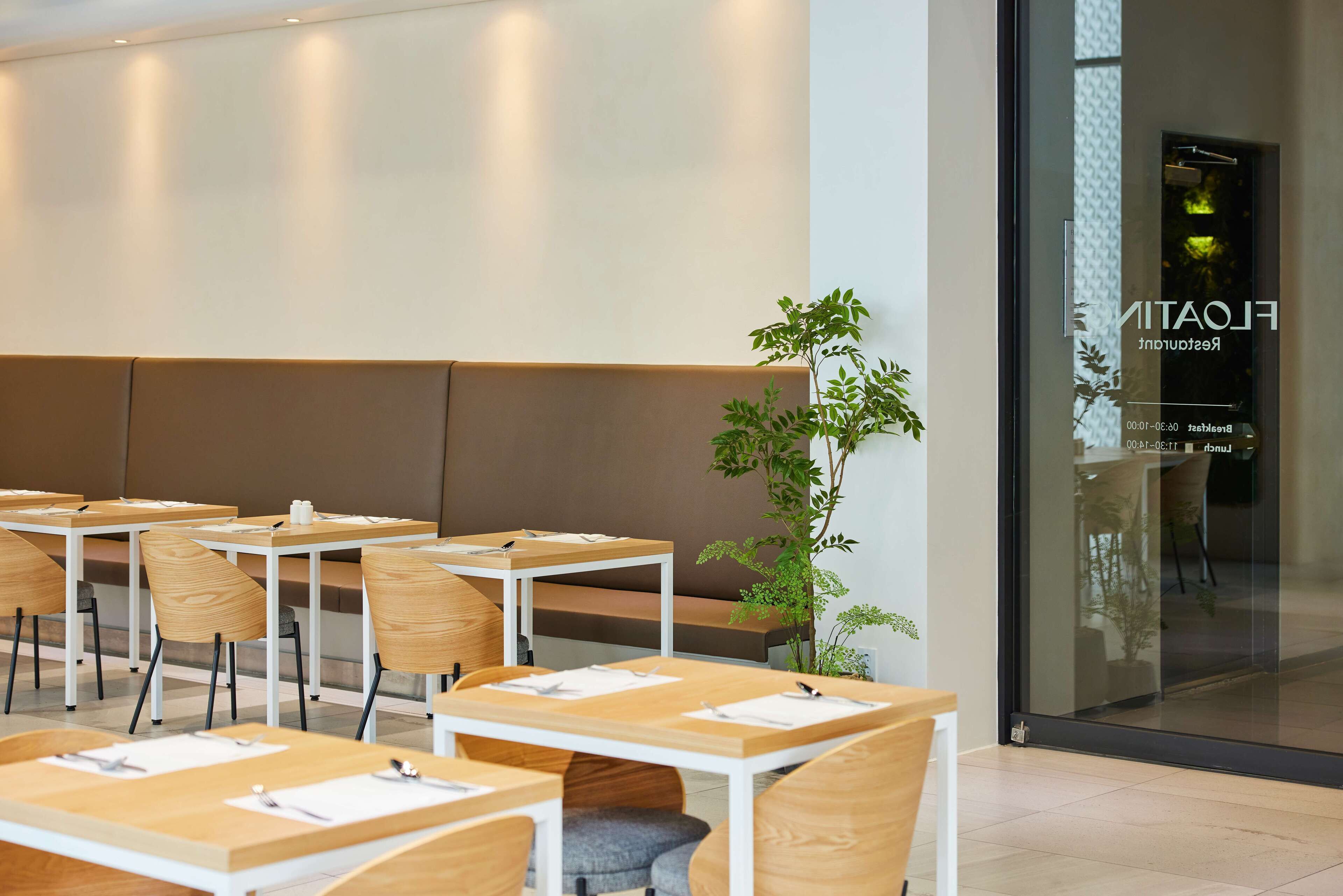 Un hall d’hôtel moderne avec des tables et des chaises en bois, une grande plante en pot et un mur de verre sur lequel est apposée une enseigne.