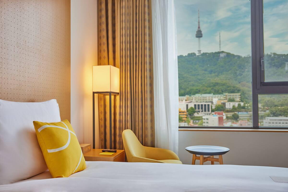 Hotelkamer met een bed, een stoel, een tafeltje, een lamp en uitzicht op een toren en stadsbeeld.