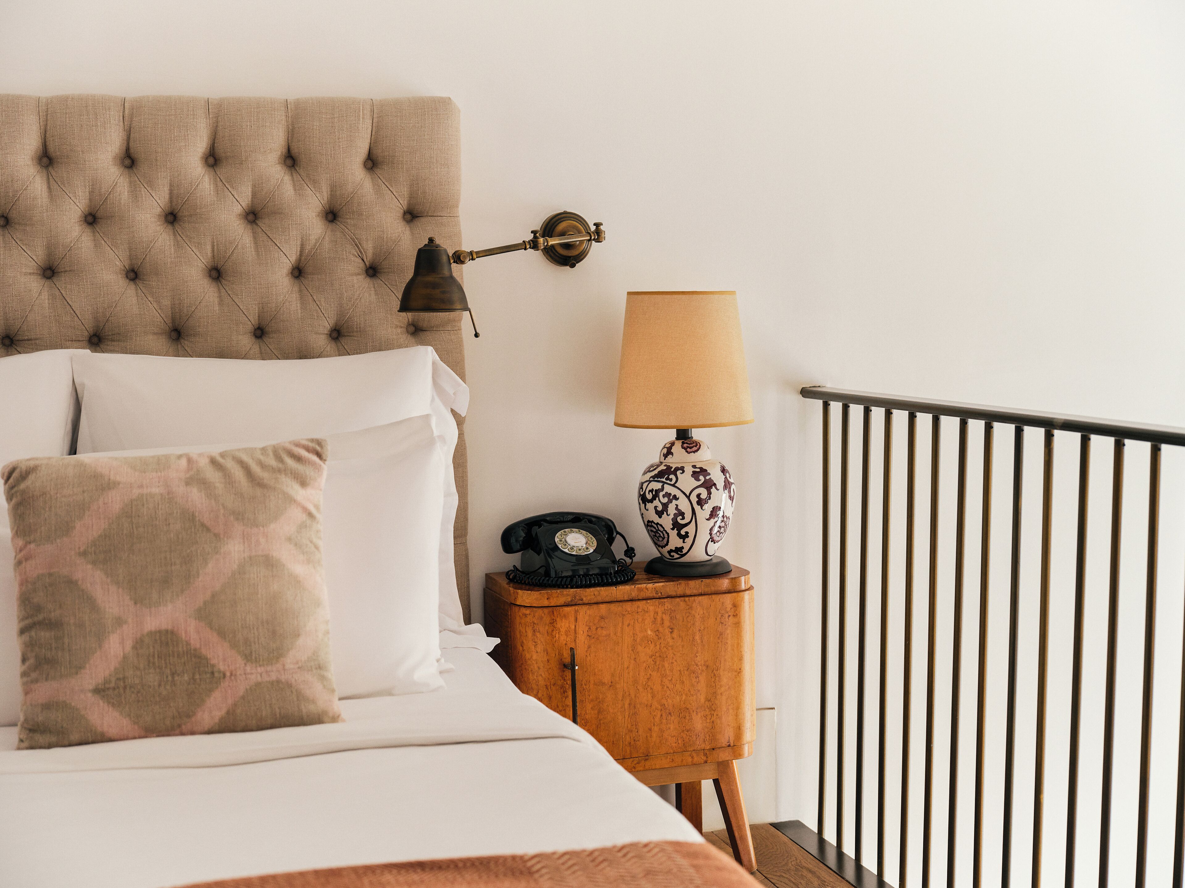 A neatly made bed with a tufted headboard, a bedside table with a lamp and a rotary phone, and a railing.