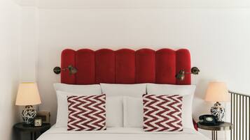 Un lit avec une tête de lit rembourrée de tissu rouge, une literie blanche et deux coussins à motifs. De chaque côté du lit, on trouve des tables de chevet avec des lampes et une horloge.