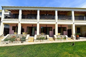 A two-story building with a balcony on the upper floor, a stone pathway leading to the entrance, and outdoor seating. - Medusa Luxury Apartments (Corfu)