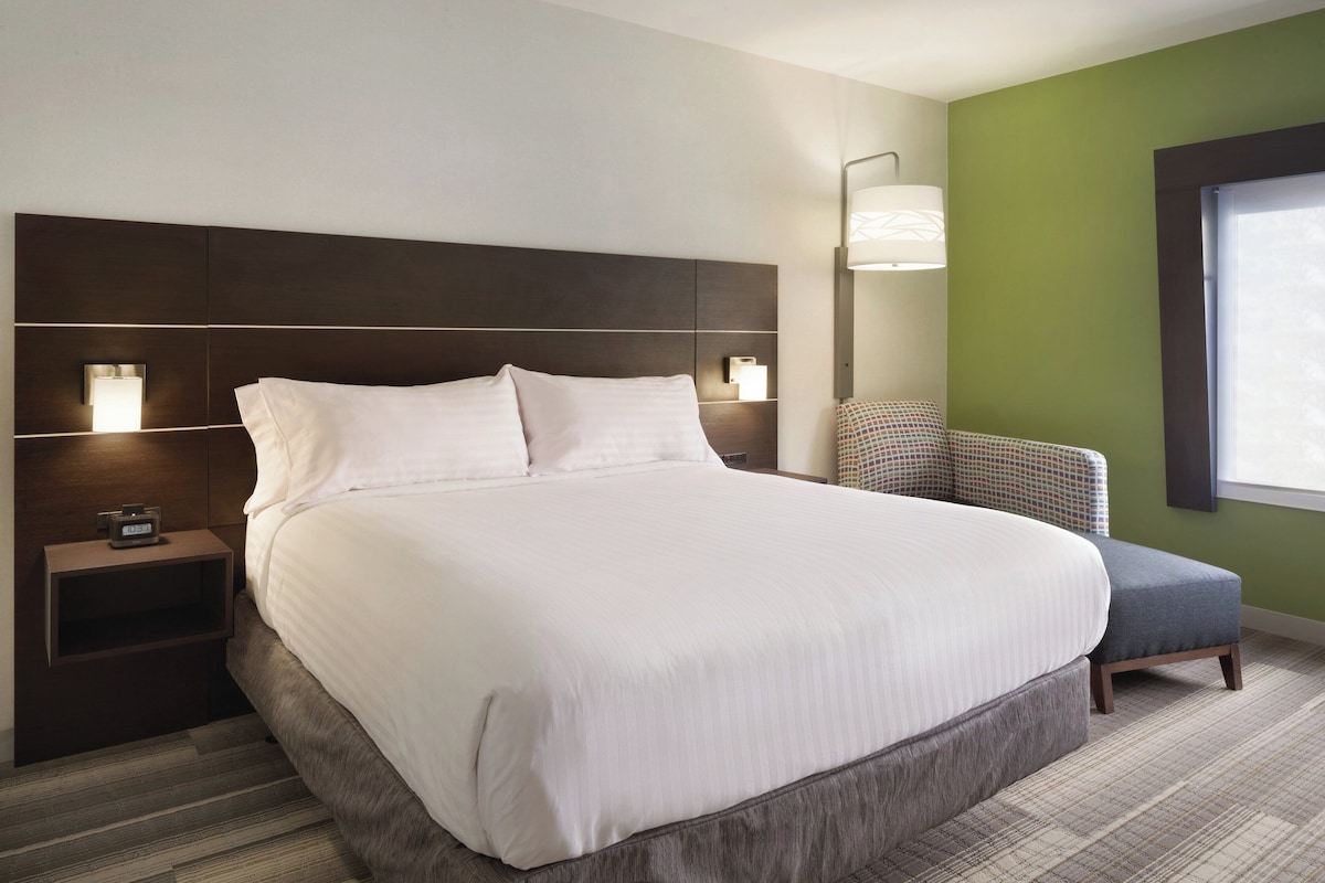 A neatly made bed with a headboard, bedside tables with lamps, a chair, and a window with blinds.