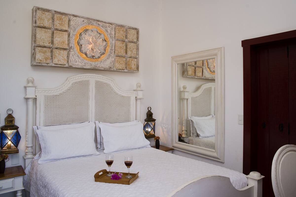 A neatly made bed with white linens and pillows, a tray with two glasses of wine, and a decorative mirror reflecting the room's interior.