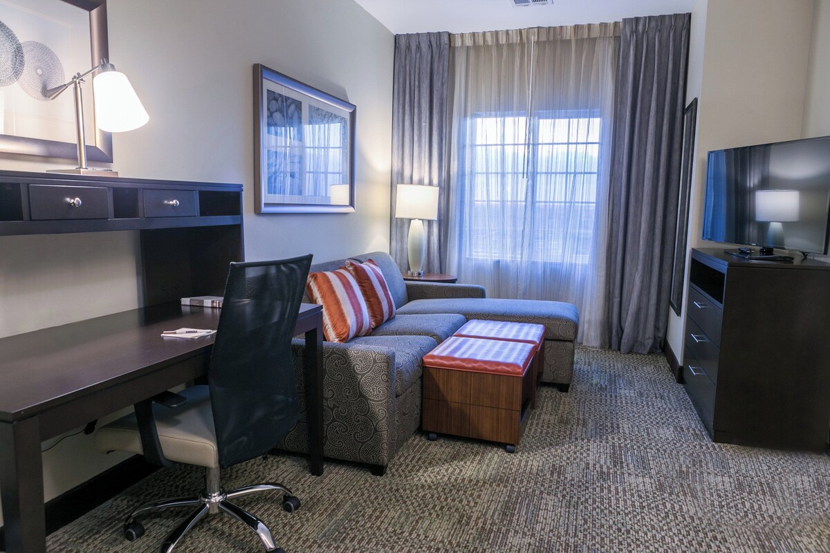 A hotel room with a desk, chair, sofa, ottoman, TV, lamp, and curtains.