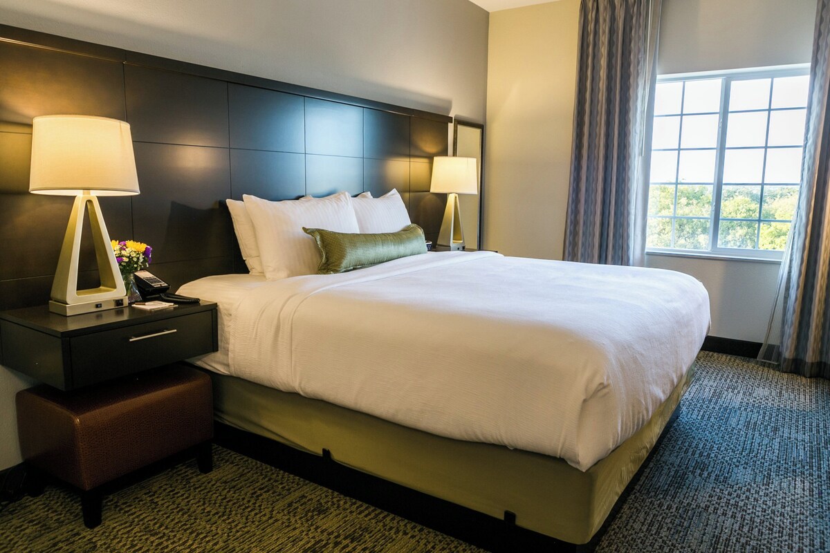 A hotel room with a large bed, two bedside tables with lamps, a small bench, and a window with curtains.