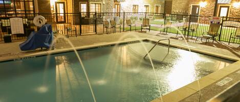 An indoor swimming pool with a covered slide, surrounded by a fence and chairs, with a building in the background.