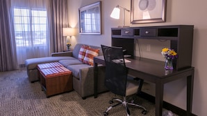 A hotel room with a sofa, a desk, a chair, a lamp, a vase with flowers, and a framed picture on the wall. - Staybridge Suites Austin North - Parmer Lane by IHG (Austin)