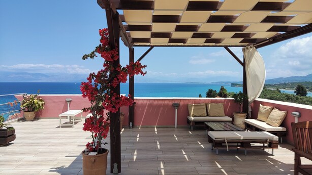 A covered outdoor seating area with a view of the sea, wooden flooring, and potted plants. - Villa Bambas (Corfu)