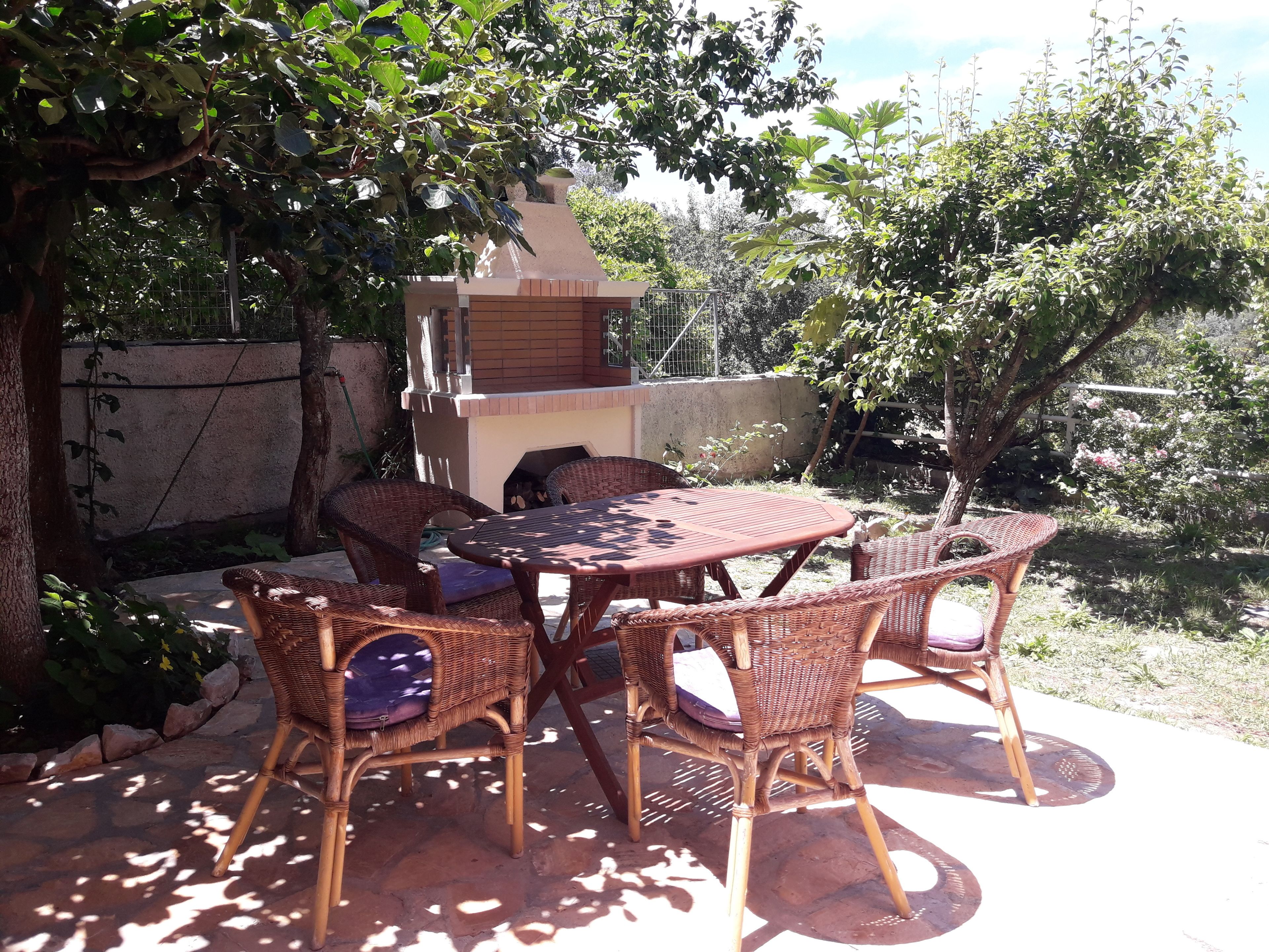 An outdoor patio with wicker furniture and a brick table, surrounded by greenery and a stone wall.