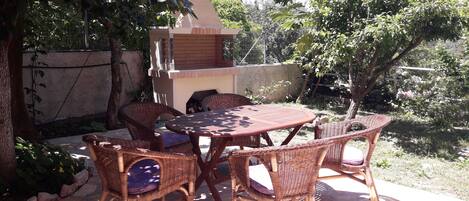 An outdoor patio with wicker furniture and a brick table, surrounded by greenery and a stone wall.