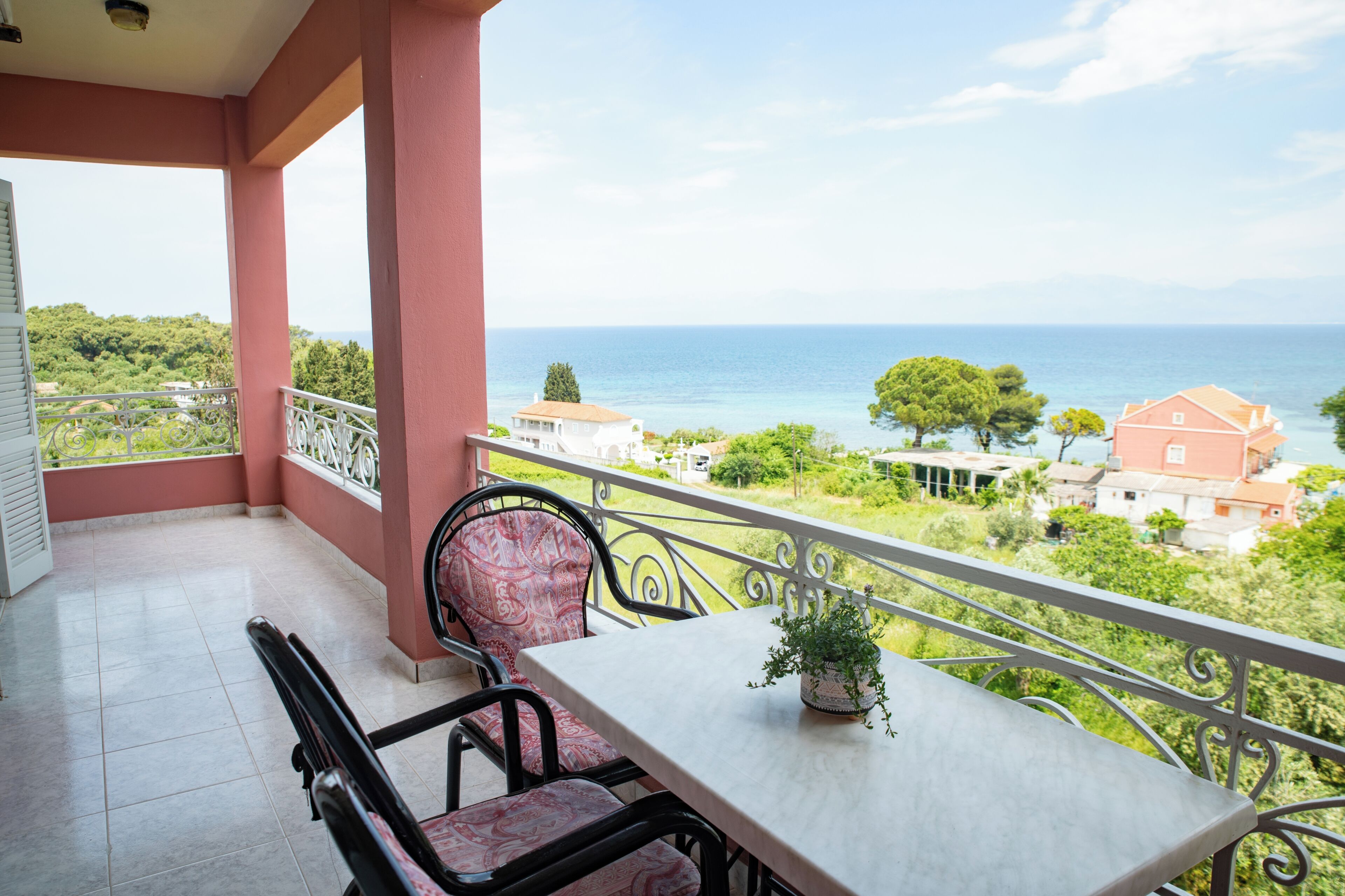 A balcony with a table and chairs, a potted plant, and a view of the sea and greenery.