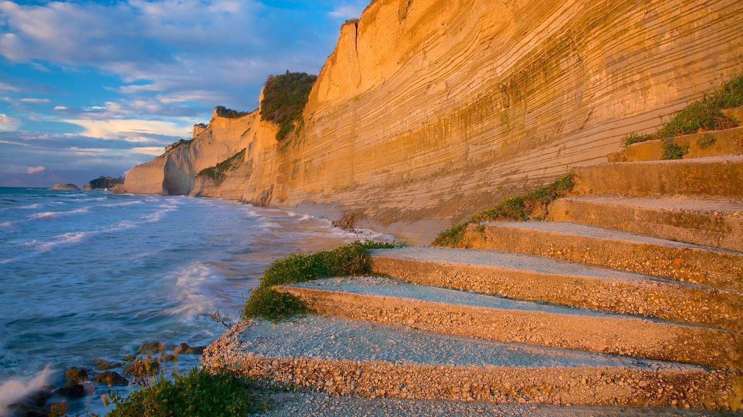 Eine Küstenstiege führt zum Strand hinab, mit einer Klippe im Hintergrund und dem Meer links.