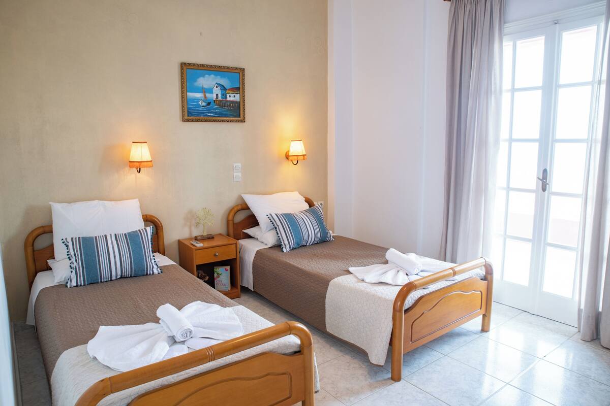 A hotel room with two wooden beds, each with a pillow and a blanket. There is a nightstand with a lamp between the beds. A painting hangs on the wall above the beds.