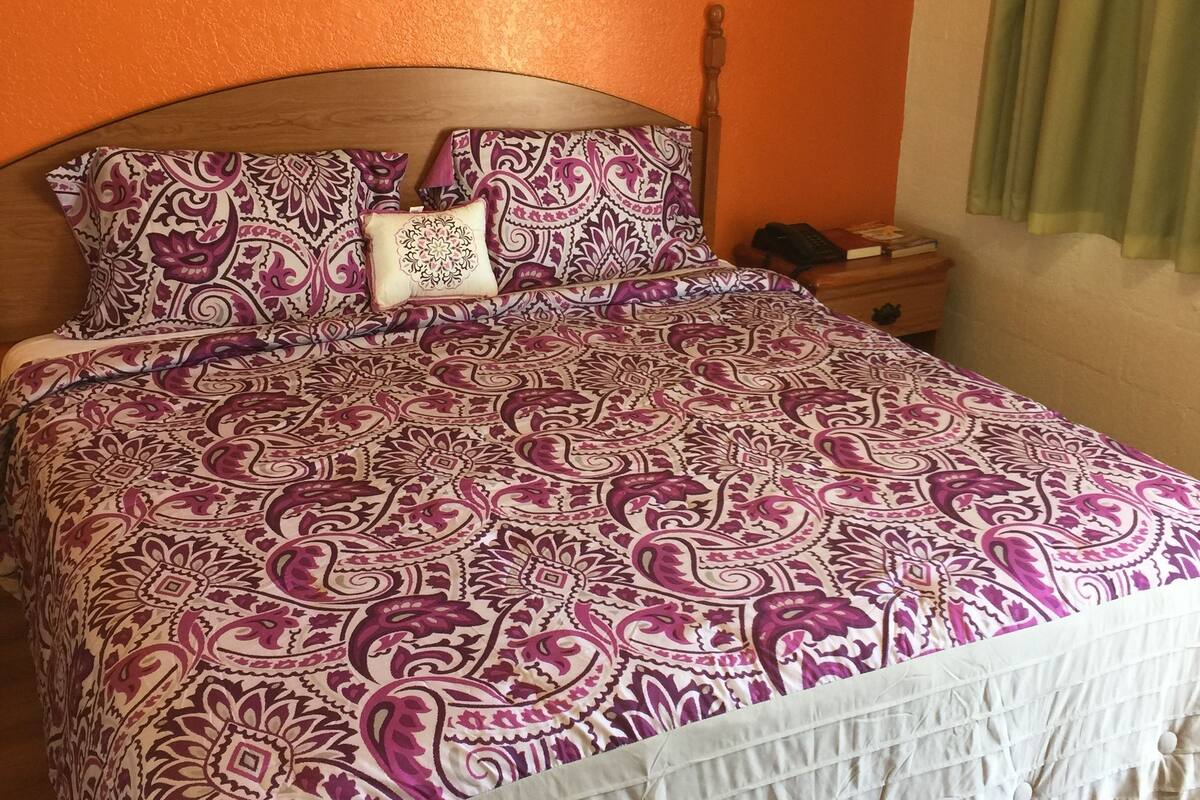 Uma cama com colcha estampada e almofadas, cabeceira de madeira, mesa de cabeceira com um livro e uma moldura na parede.