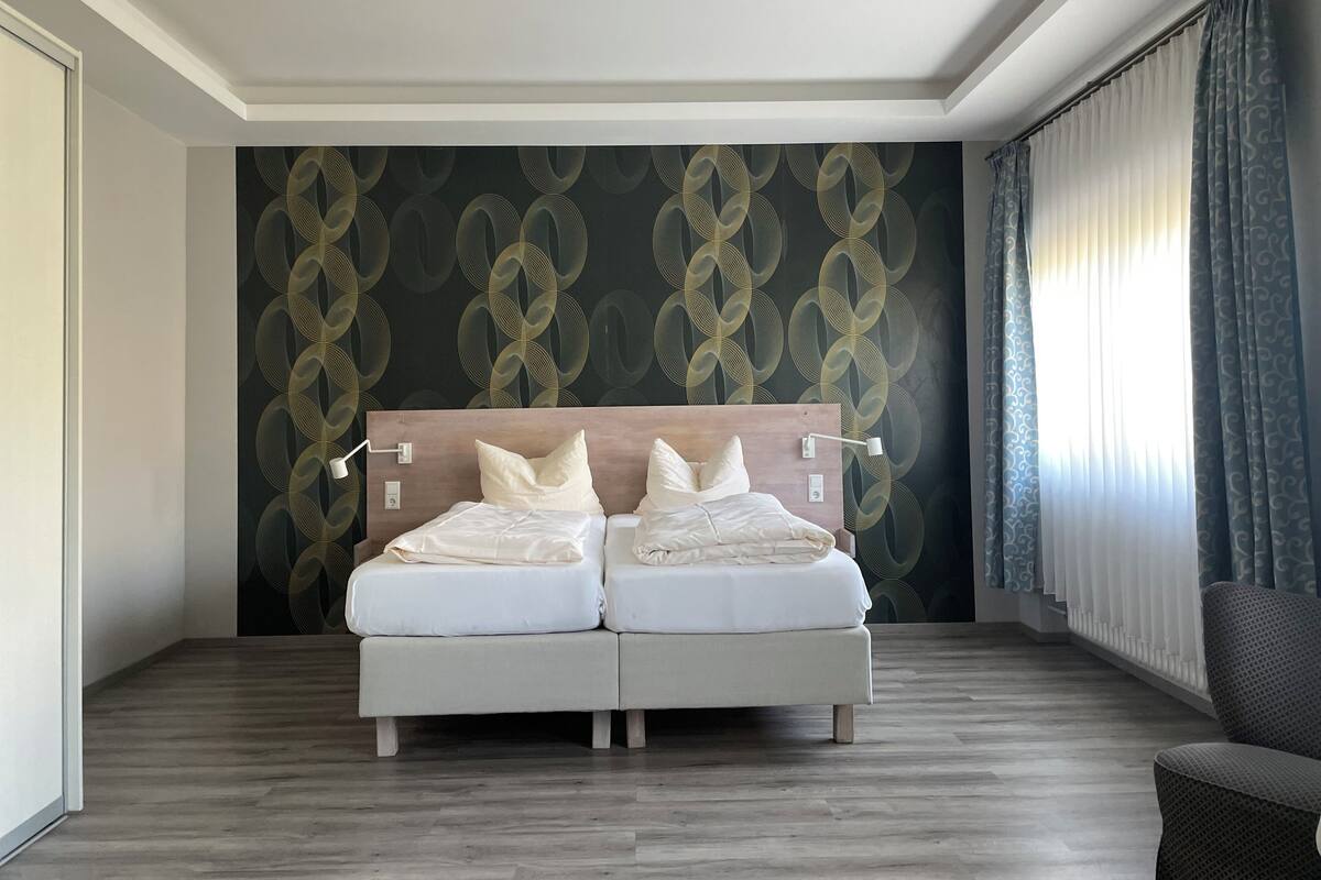 Ein Doppelbett mit weißer Bettwäsche in einem Hotelzimmer mit dunkel gemustertem Wandteppich und einem Holz-Kopfende.
