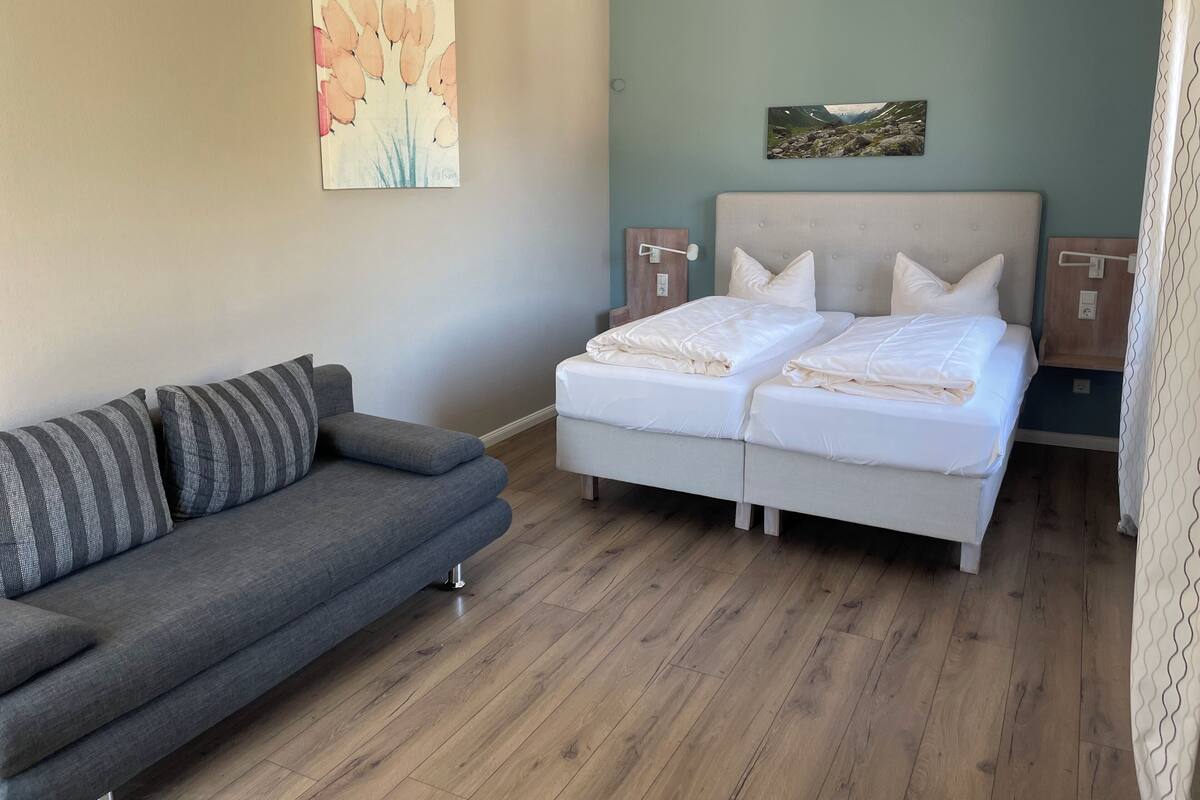 Ein Schlafzimmer mit einem Doppelbett, einem grauen Sofa, Holzboden und zwei an der Wand befestigten Nachttischen.