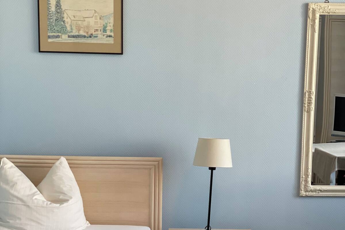 Ein ordentlich bezogenes Bett mit weißen Leinen und Kissen, ein Nachttisch mit Lampe, ein gerahmtes Bild an der Wand und ein großer Spiegel.