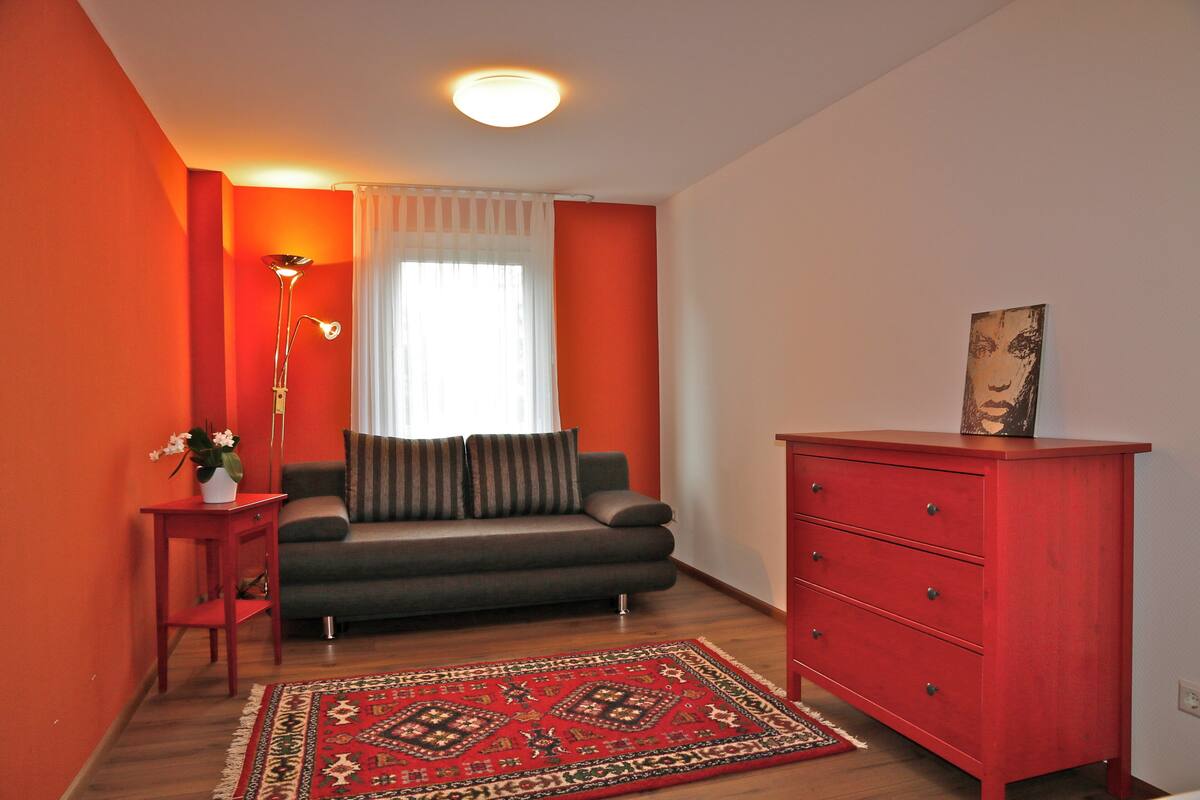 Ein Wohnzimmer mit einer Couch, einem roten Schminktisch, einem gemusterten Teppich und einer Stehlampe.