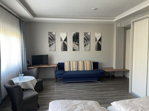 A modern hotel room with a blue sofa, a small round table with a white tablecloth, a television, and a series of black and white photographs on the wall.
