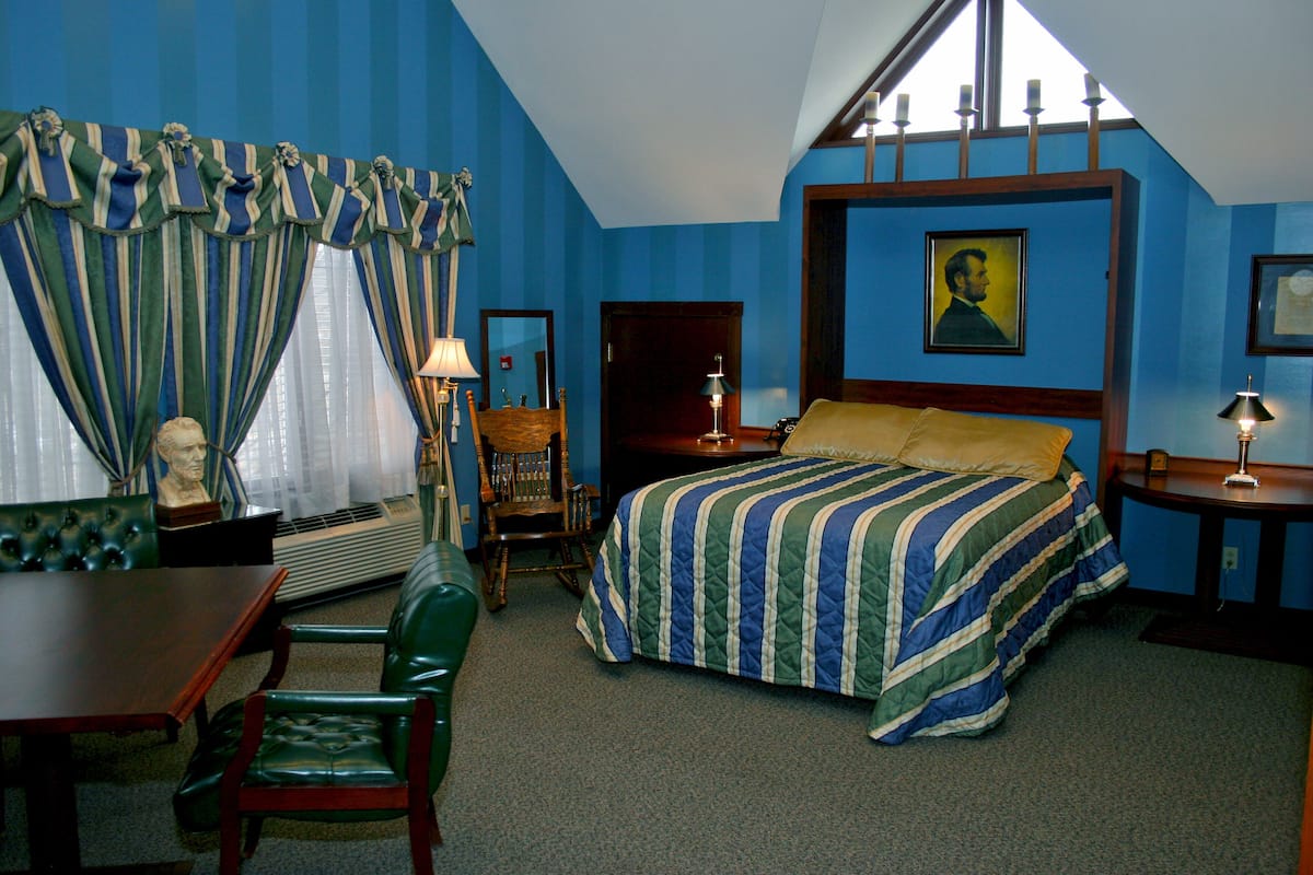 A bedroom with a bed, a desk, a chair, a lamp, and a portrait on the wall.
