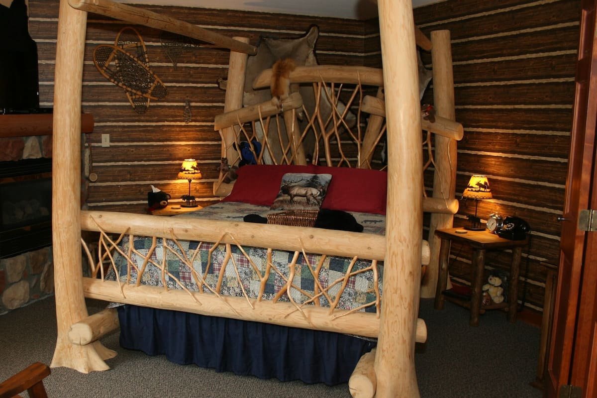 A rustic wooden bed with a woven headboard, a red pillow, and a patterned bedspread. There are two bedside lamps and a small table with a lamp.