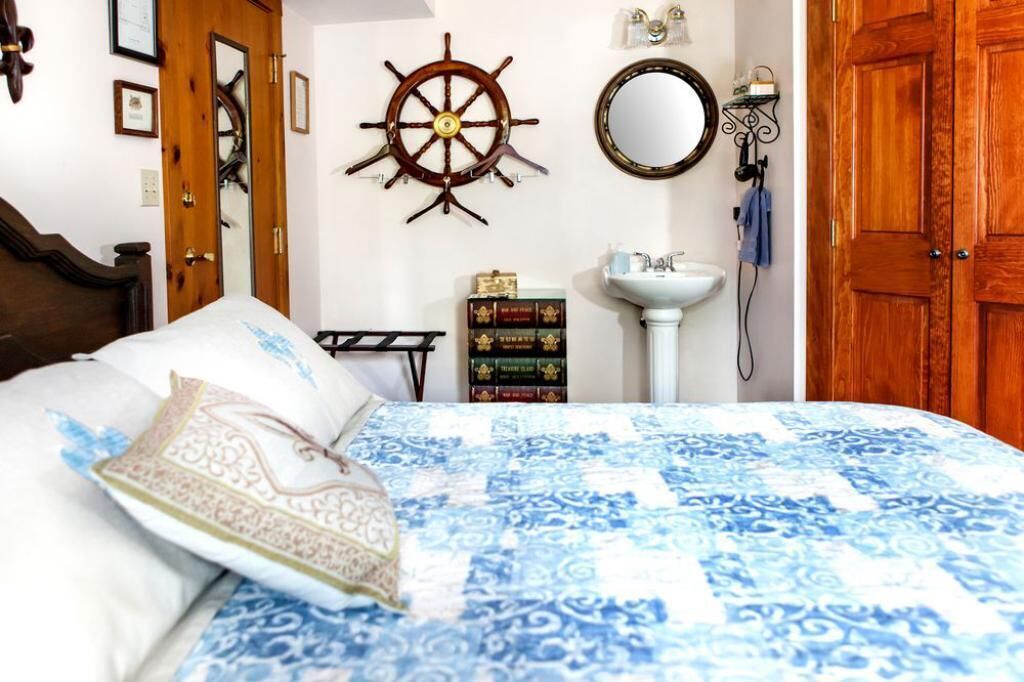 Una camera da letto con un letto, una porta in legno, una ruota di timone a muro, uno specchio, una libreria e un lavandino bianco.