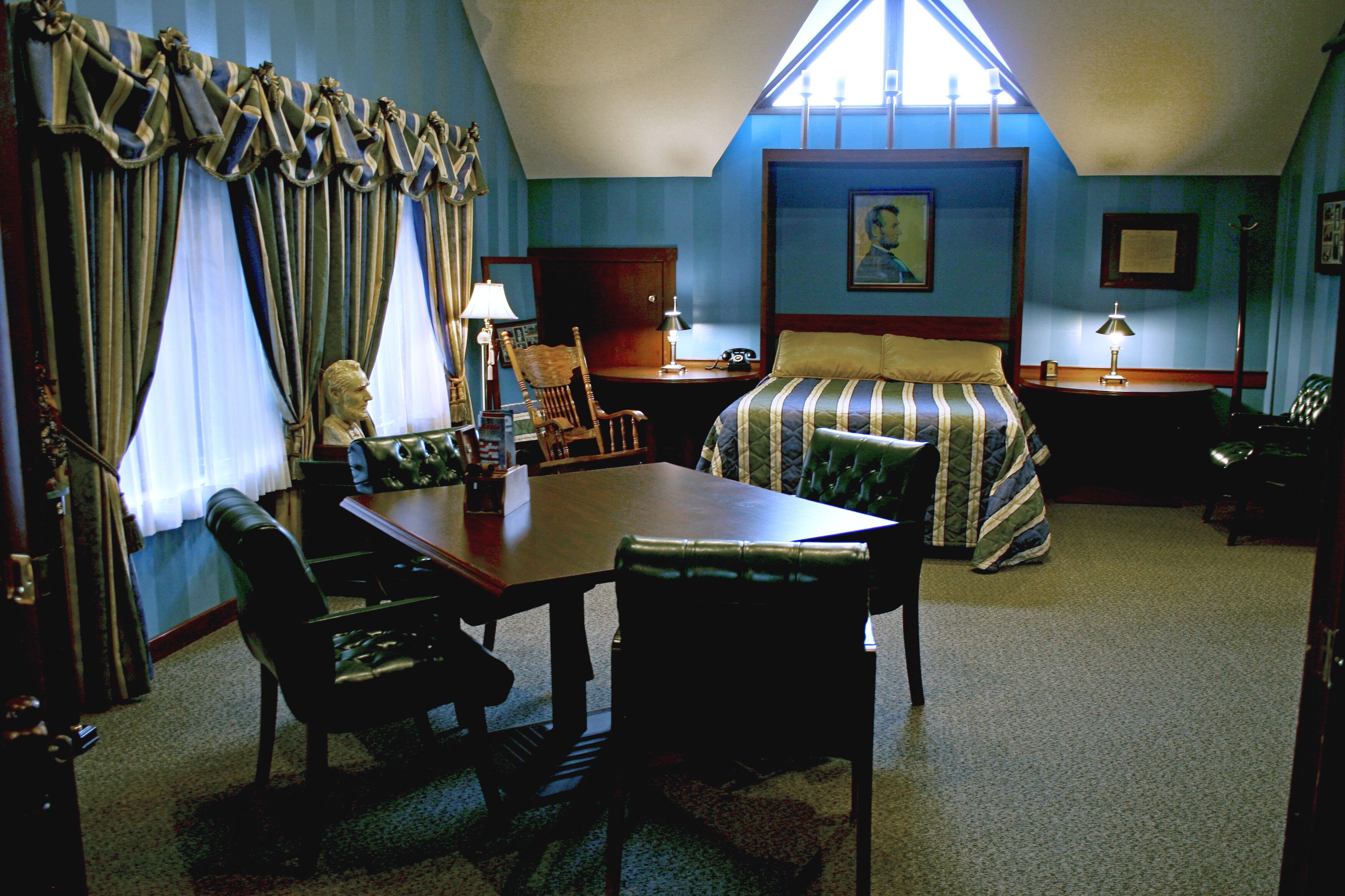 A room with a large bed, a desk, two chairs, a window with curtains, and a painting on the wall.
