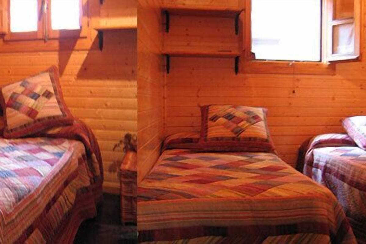 Une chambre confortable, aménagée dans le style d’un chalet, avec deux lits, chacun agrémenté d’une courtepointe matelassée et de coussins. On y trouve également un petit banc en bois et une étagère au-dessus des lits.