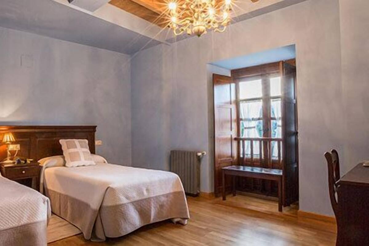 Une chambre d’hôtel avec deux lits, une tête de lit en bois, un lustre, une fenêtre et une table.