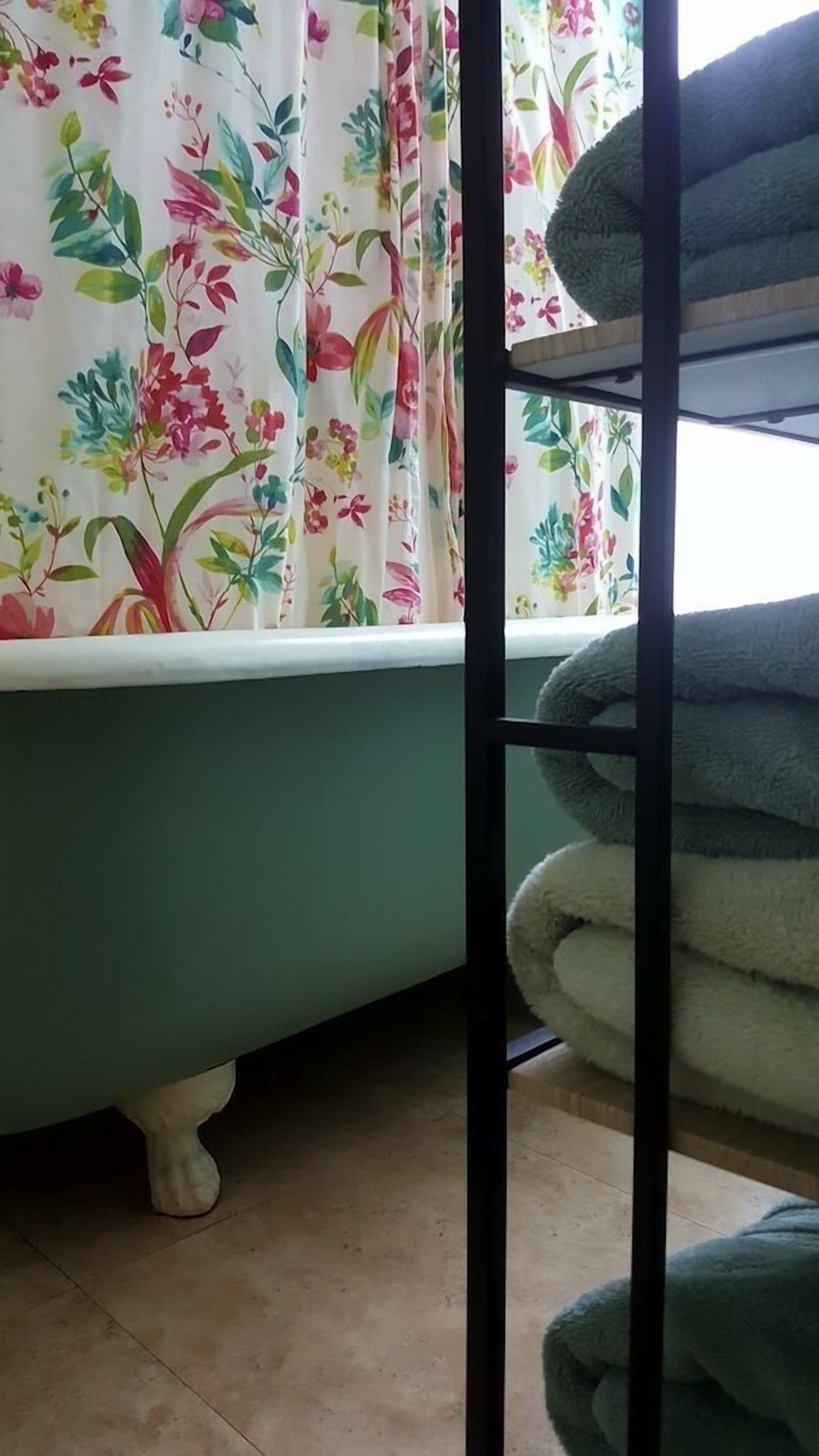 A bathroom with a green bathtub, floral curtains, and a shelf with folded towels.
