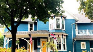 A two-story house with a blue exterior, a front porch, and a flagpole with two flags. - The Muse Gallery Guesthouse (Milwaukee)