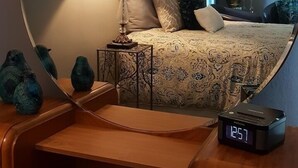 A bedroom with a round mirror, a bed with patterned bedding, a bedside table with a lamp, and a digital clock. - The Muse Gallery Guesthouse (Milwaukee)