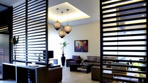 Una sala de estar moderna con un sofá modular, una mesa de centro y un televisor. Hay dos ventanas grandes con persianas, una planta en maceta y un cuadro en la pared.