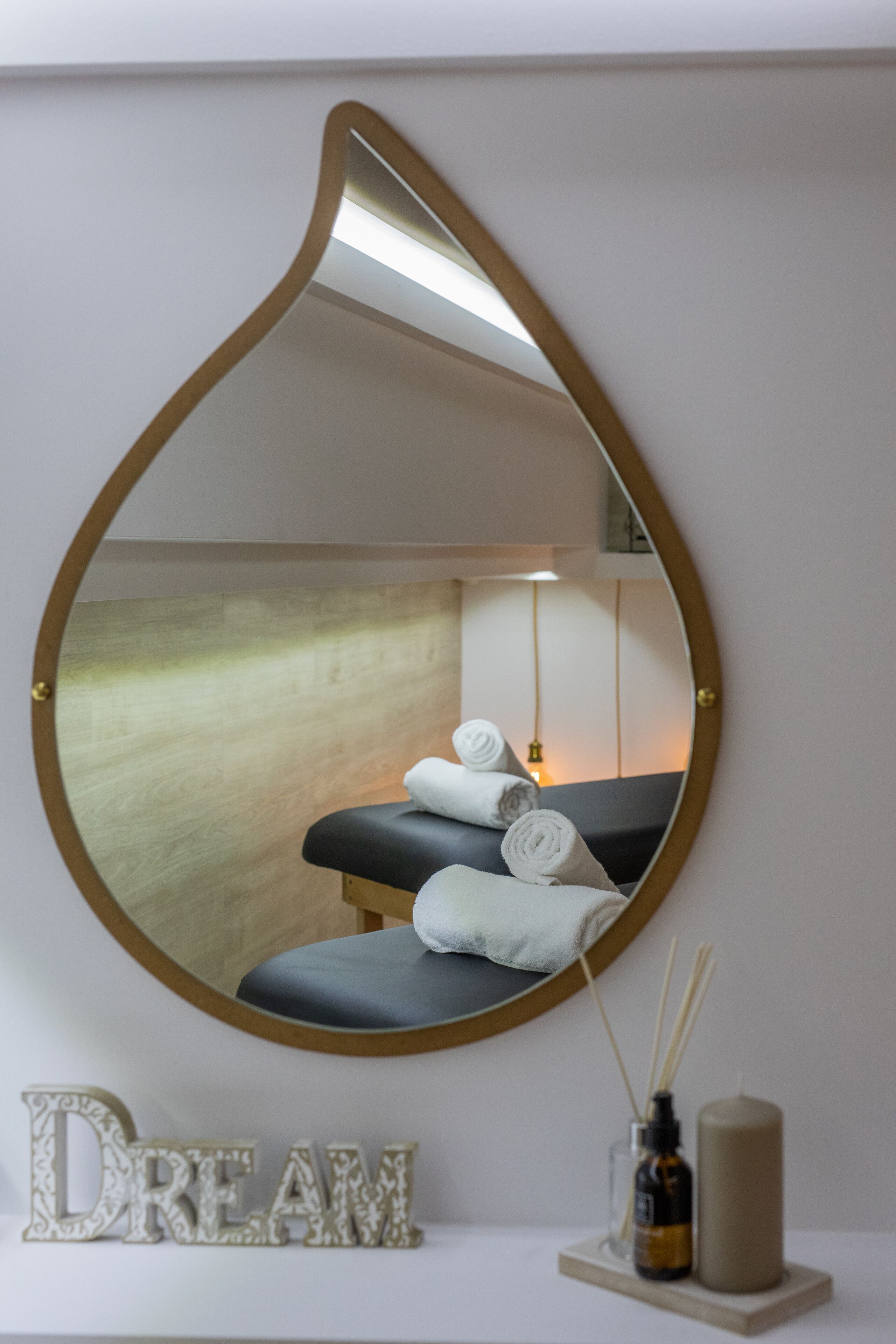 A mirror with a unique shape reflecting a spa room, a 'DREAM' sign, and a diffuser with reed sticks.