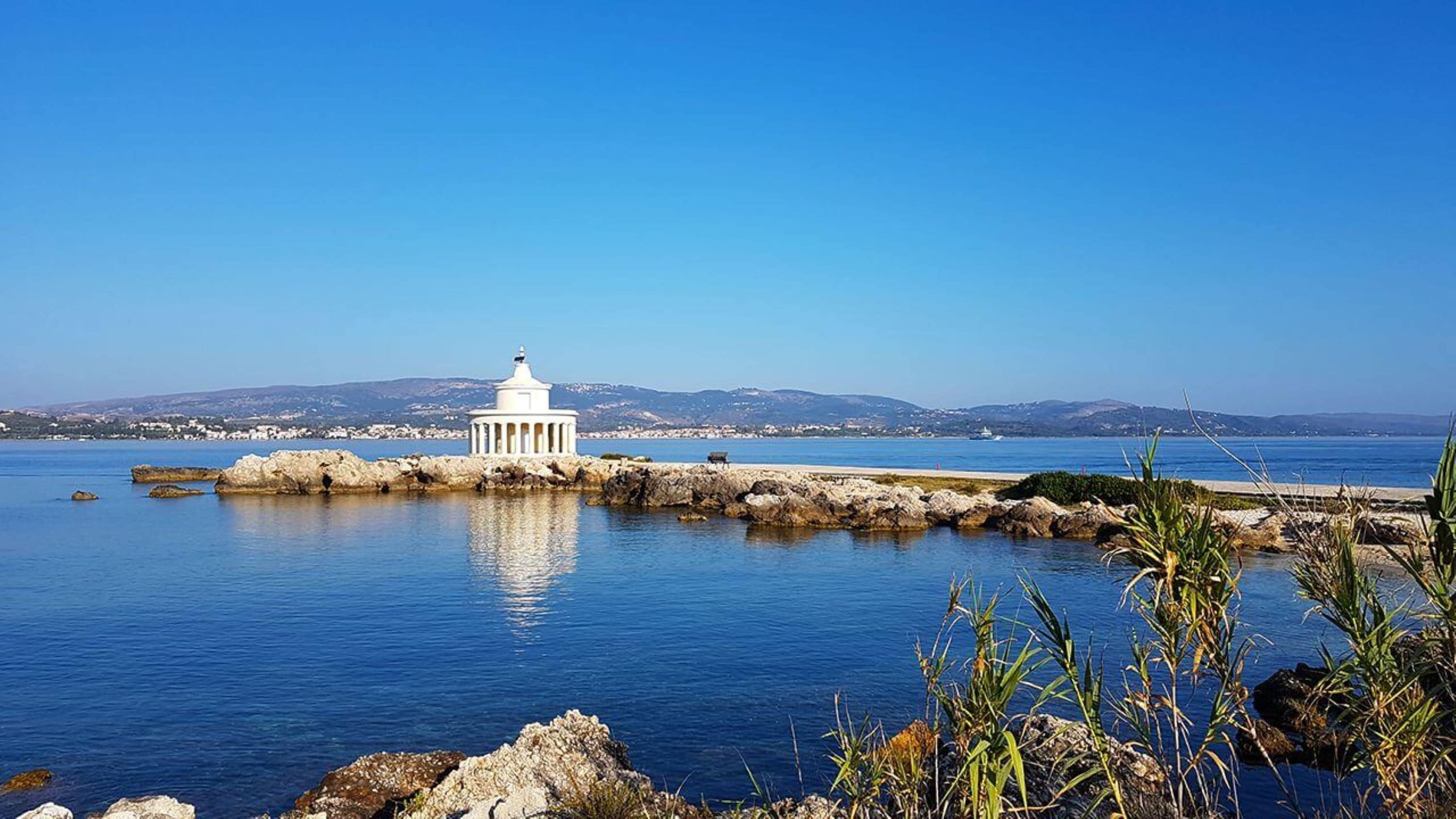 A coastal scene with a white building near the water, rocky shoreline, and distant hills.