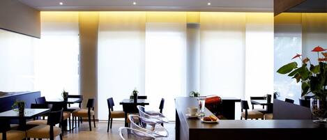 A modern restaurant with high tables and clear chairs, large windows, and a warm ceiling light.