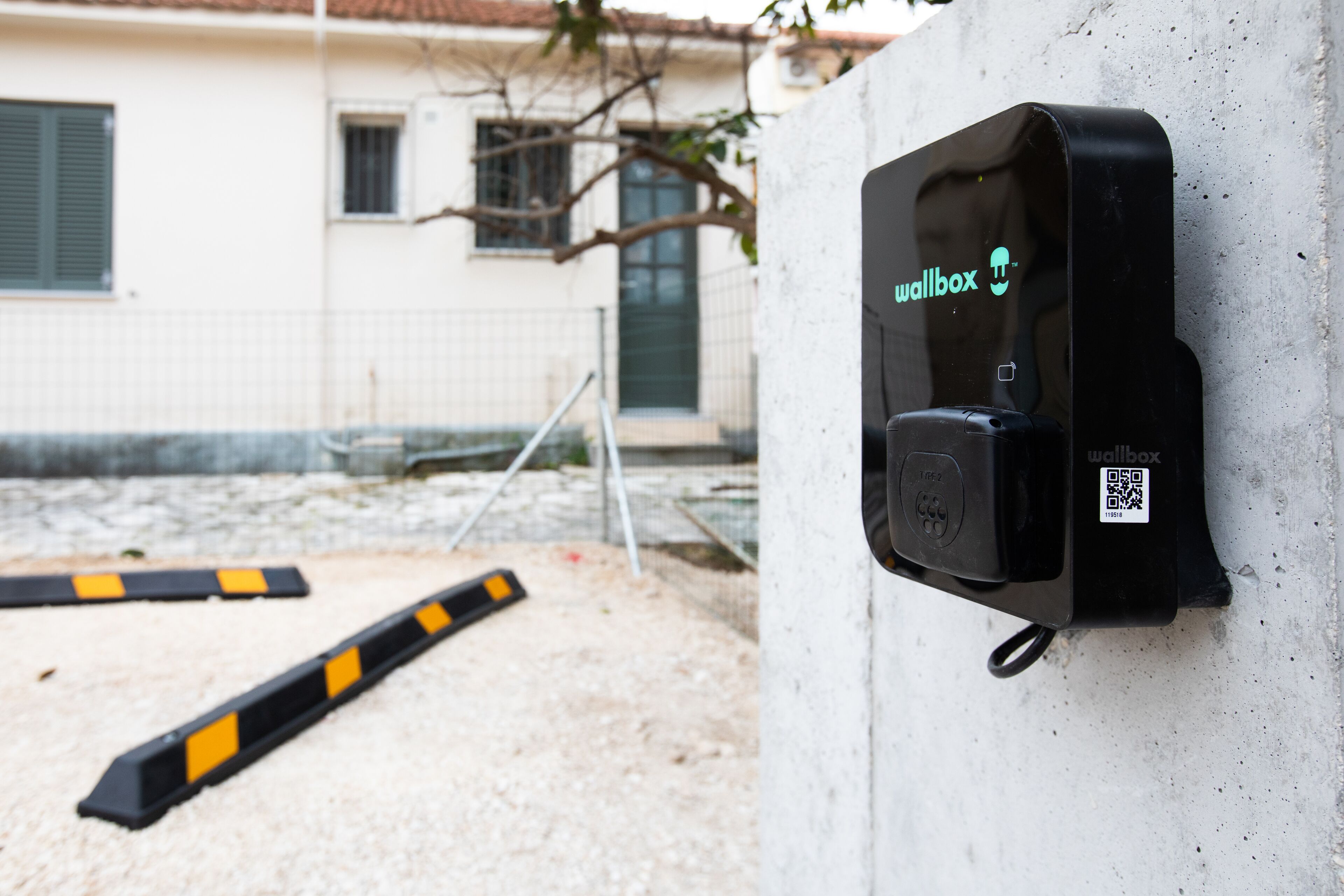 A wall-mounted wallbox with a QR code, a metal barrier, and a building in the background.