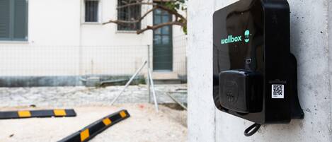 Un punto de carga mural con un código QR, una barrera metálica y un edificio al fondo.