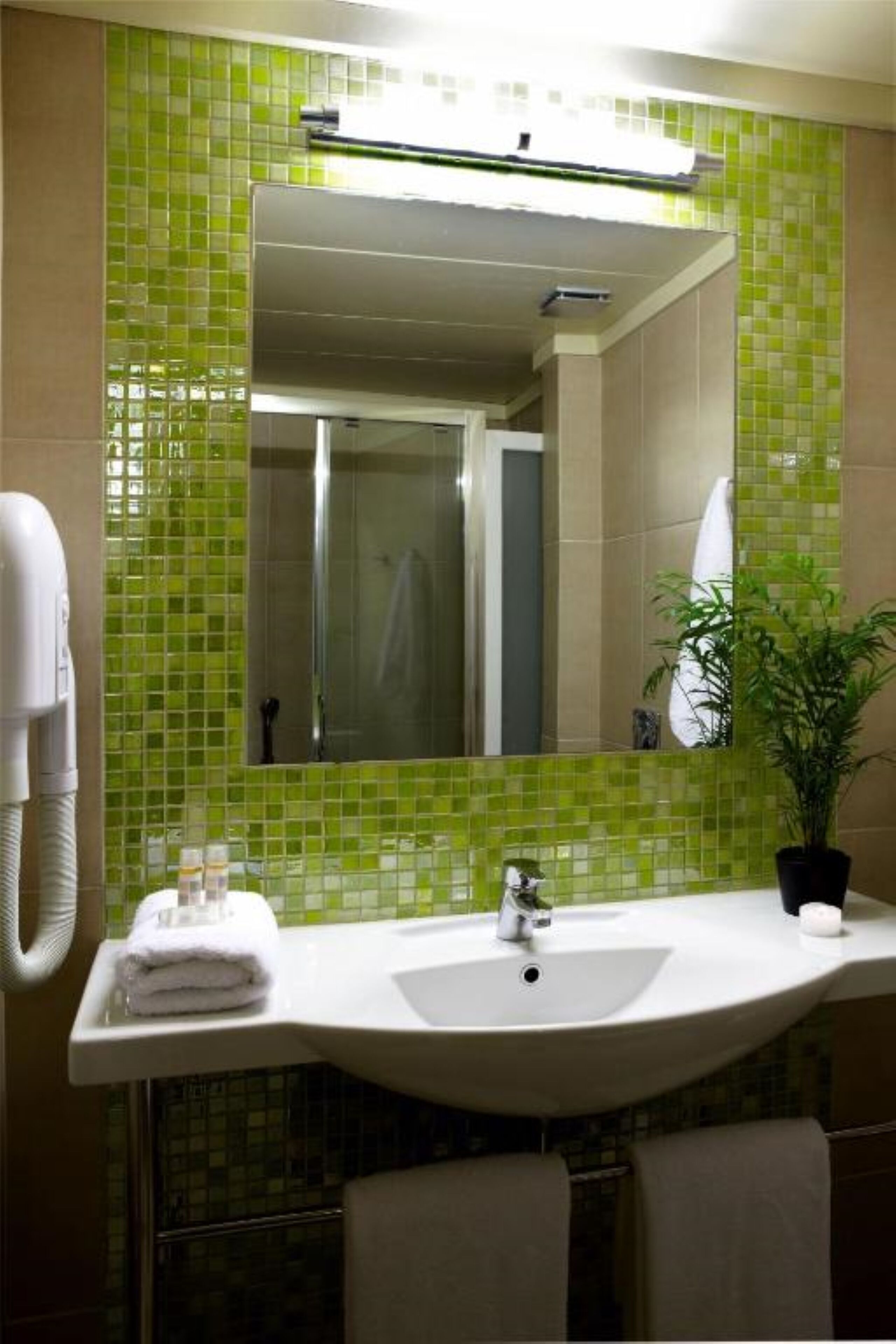 Un baño con pared de azulejos verdes, un lavamanos blanco, un espejo y una planta en maceta.