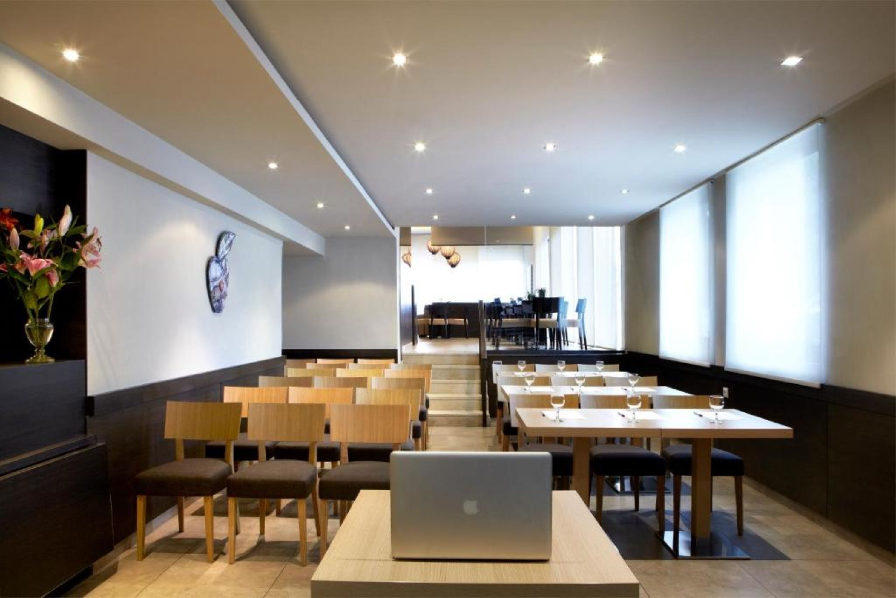 Un espace repas moderne avec des tables et des chaises en bois, un ordinateur portable posé sur l’une des tables et un vase de fleurs sur la gauche.