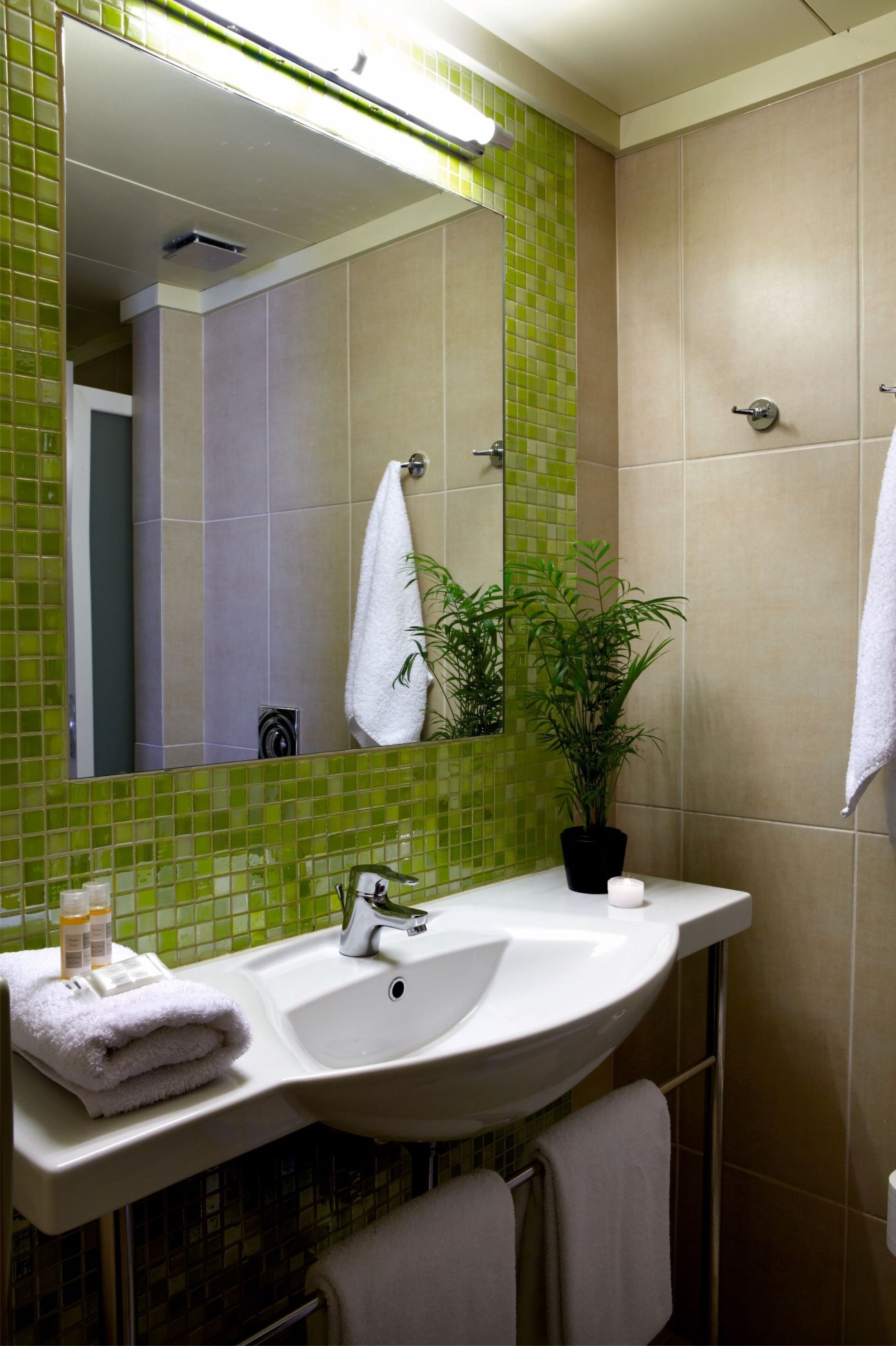 A bathroom with a green-tiled wall, a white sink, a mirror, a potted plant, and two towels.