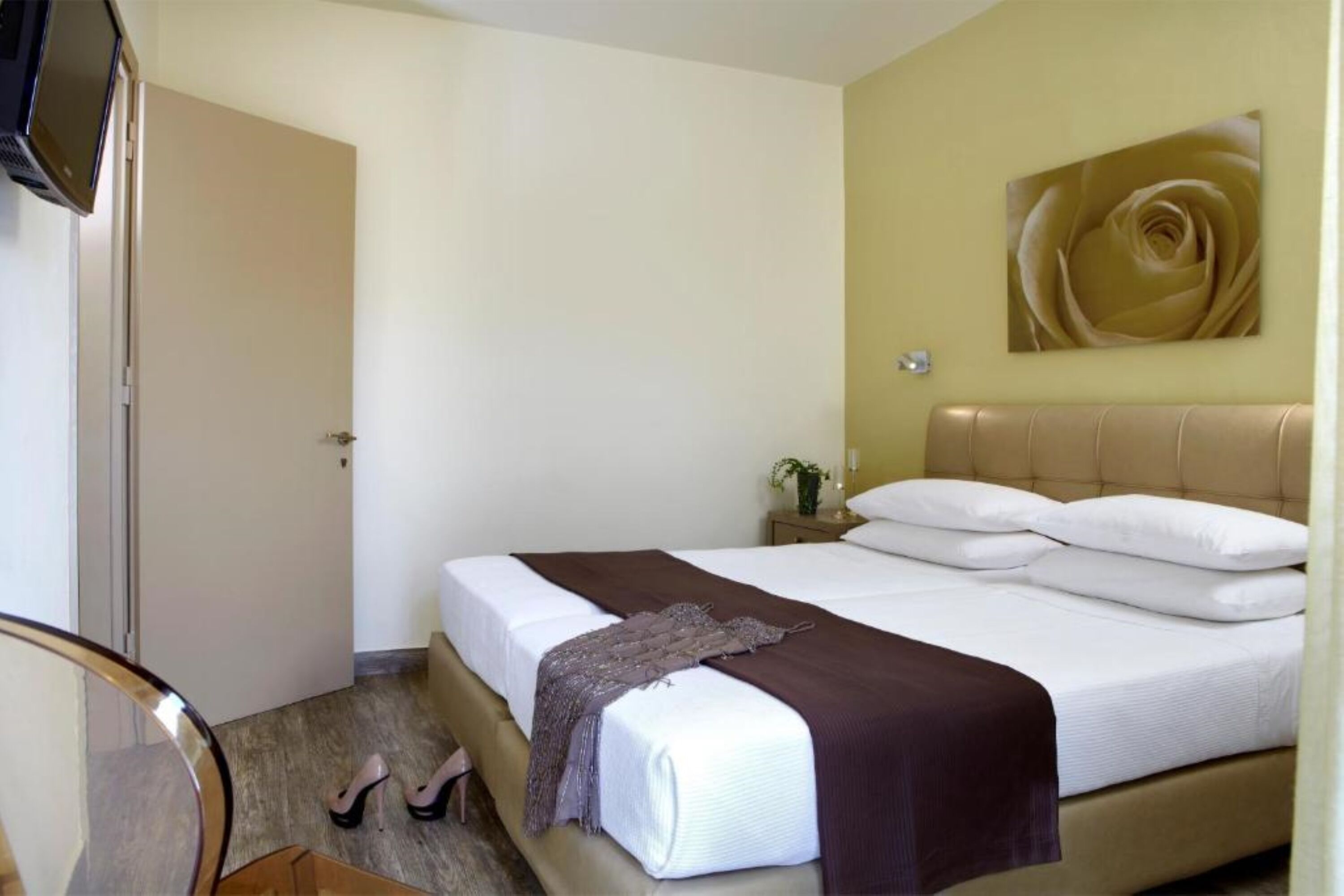 Quarto com cama, mesa de cabeceira com abajur, televisão na parede e uma porta.