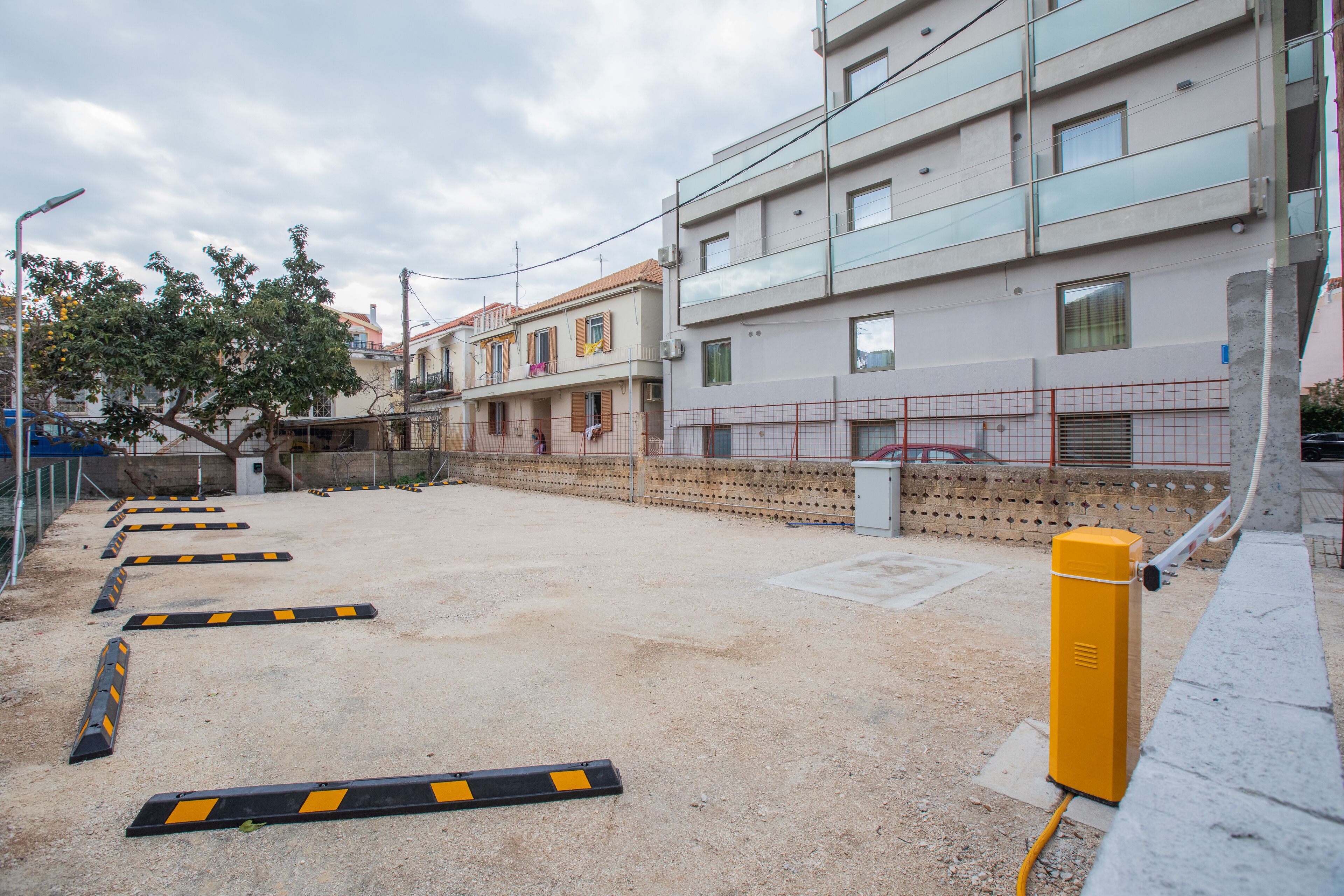 A parking lot with a yellow electric vehicle charging station, a tree, and a row of buildings in the background.