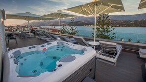 Un jacuzzi en una terraza con sillones reclinables y sombrillas, con vistas a un lago y montañas.