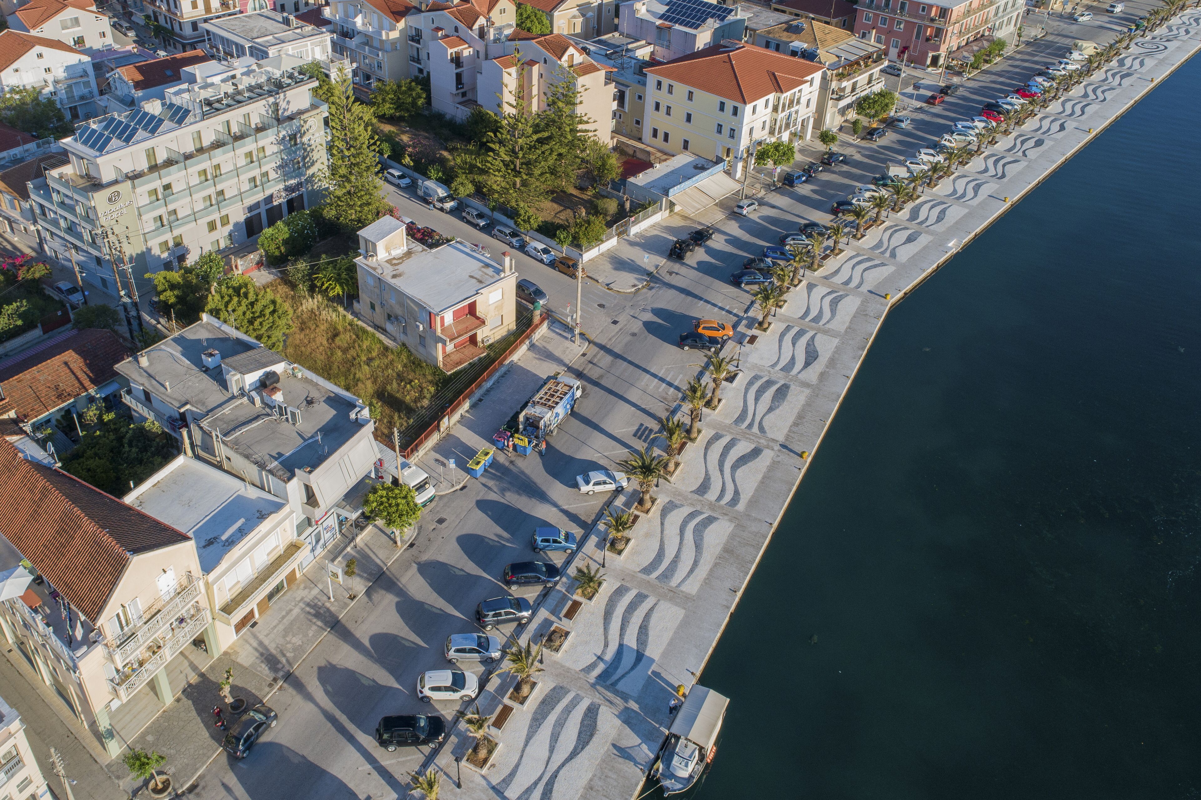 An aerial view of a coastal urban area with buildings, parked cars, and a waterfront promenade.