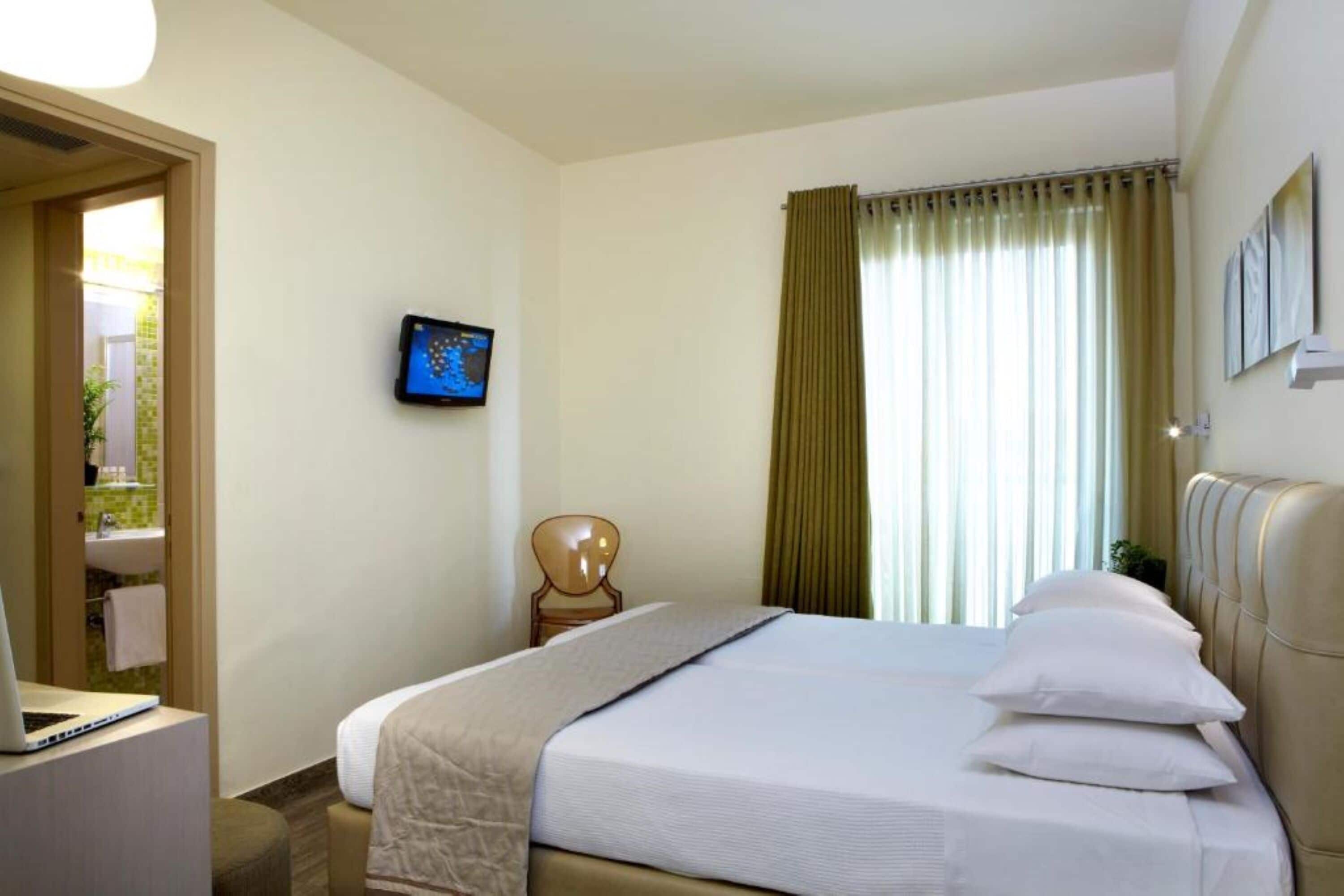 Une chambre d’hôtel avec un lit, un bureau avec un ordinateur portable, une chaise, un téléviseur fixé au mur et une fenêtre avec des rideaux.