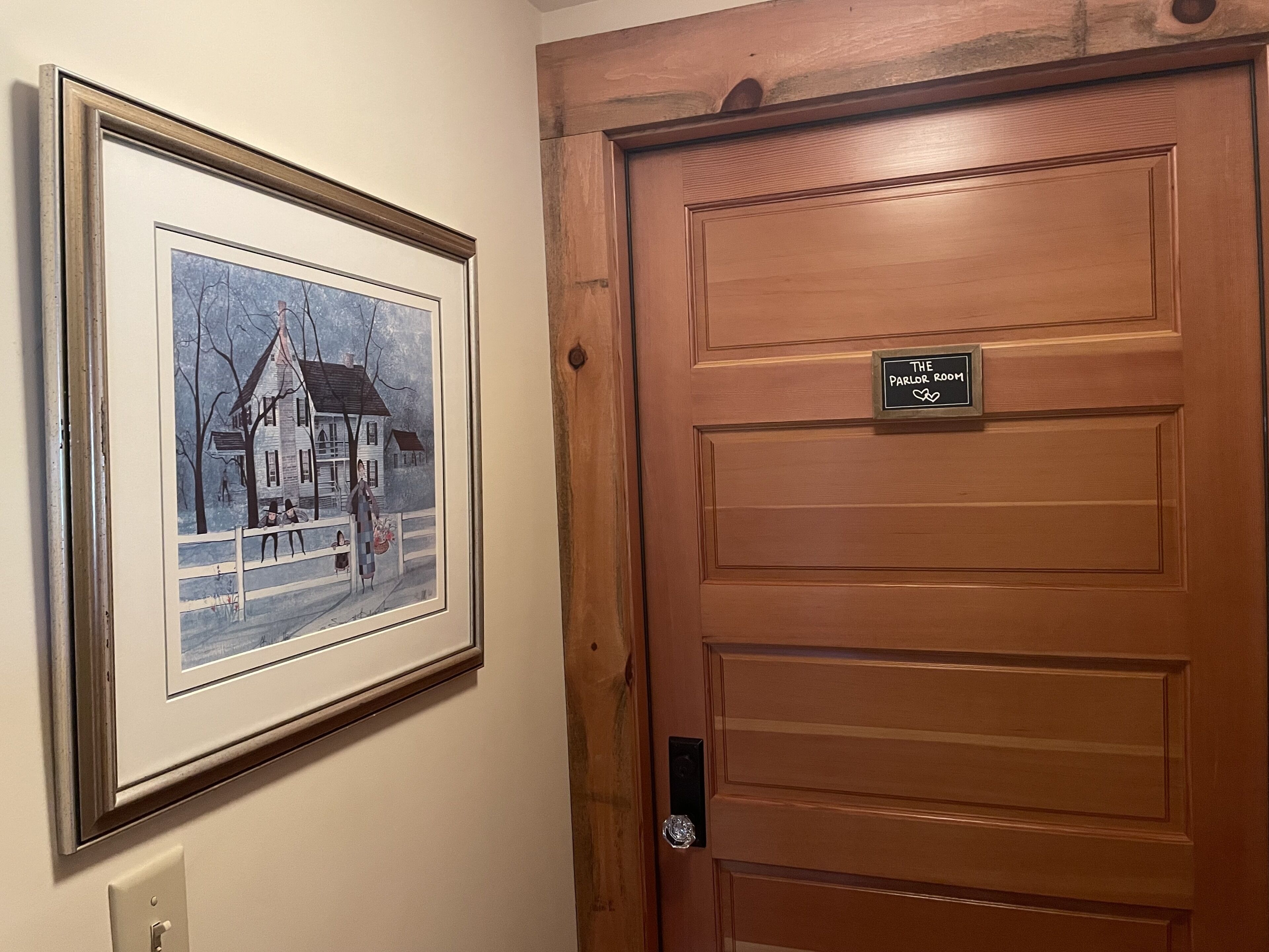 Une porte en bois sur laquelle est apposée une plaque indiquant « The Parlor Room », et un tableau encadré représentant une maison avec une personne debout près d’une clôture.